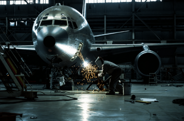Technician welding metal on an airplane nose inside a maintenance hangar.
